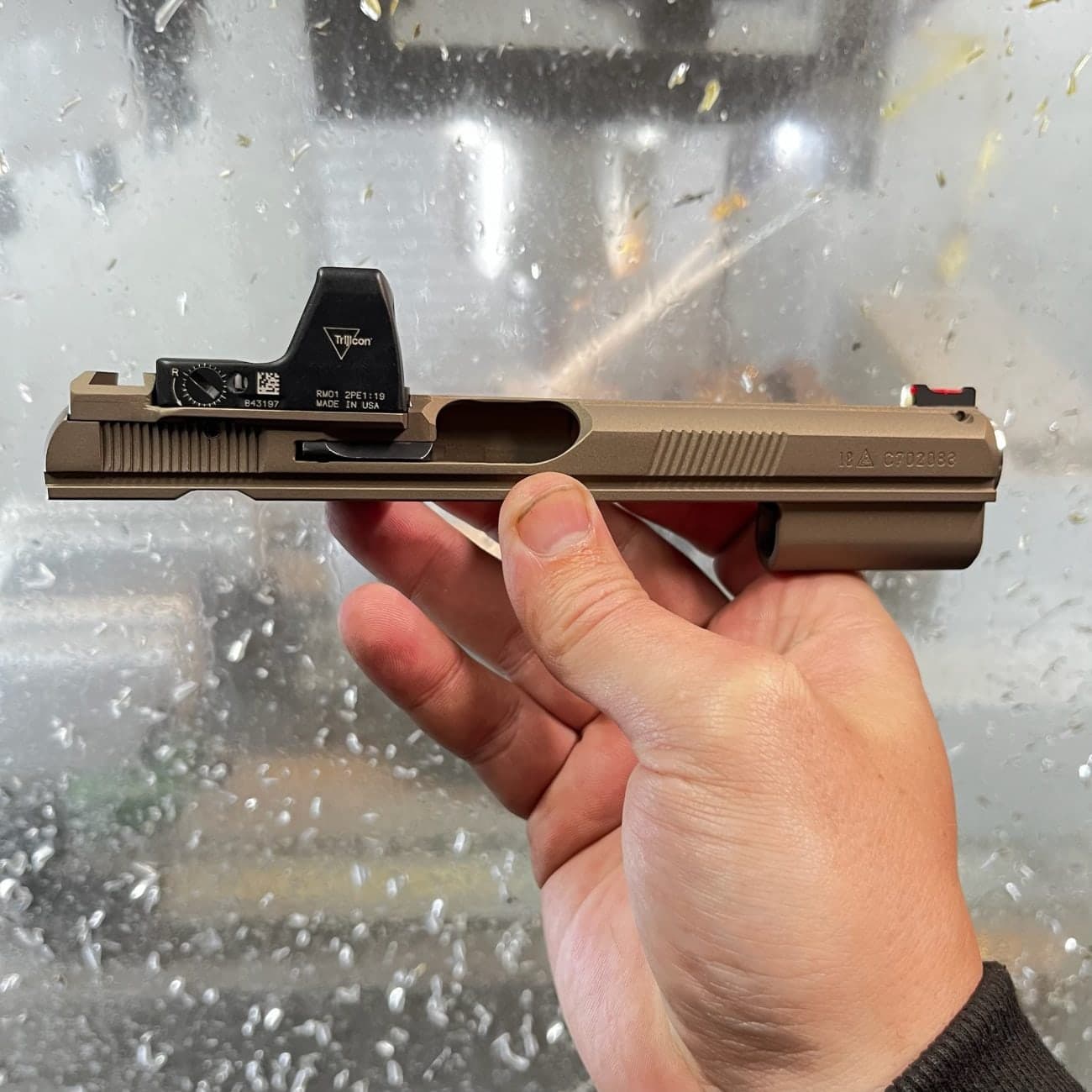Hand holding an FDE slide fitted with a red dot, in front of a coolant-spattered machine window