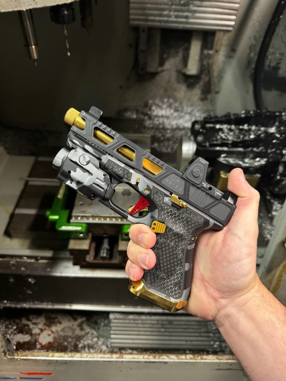 Hand holding a camo-finished pistol with gold accents inside the mill, aluminum chips on the machine bed
